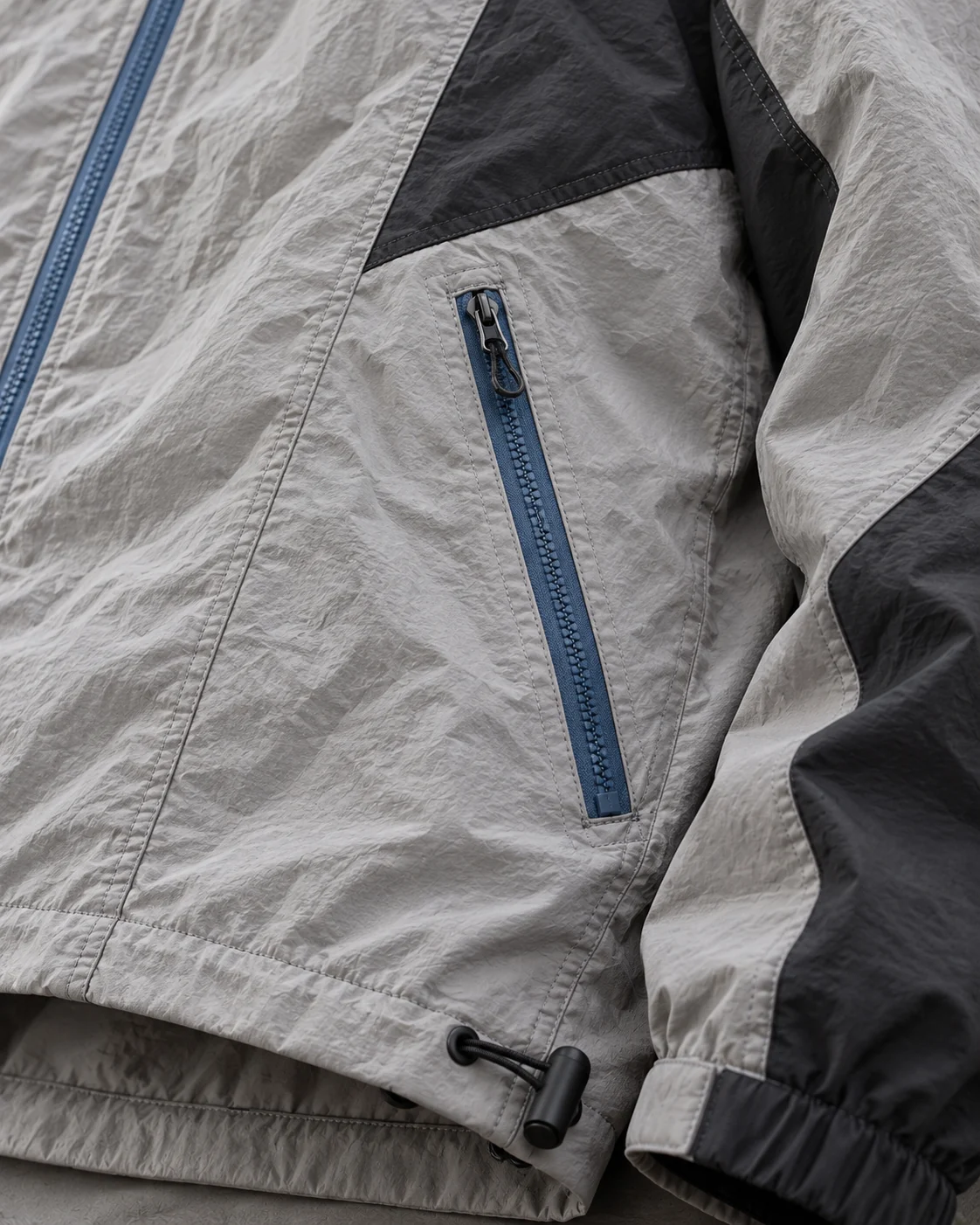 Close-up of fabric and signature construction — Hooded Shell Jacket in stone grey / charcoal / cobalt, AI-generated design concept