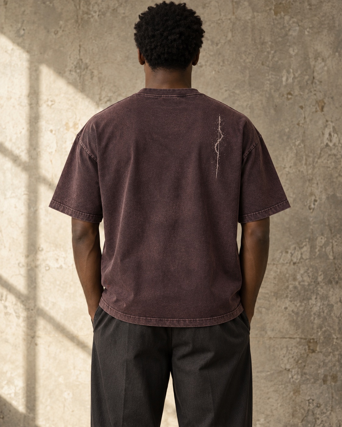 Back view of the same short-haired model in the aubergine vintage-finish T-shirt with a small broken chalk trace high on the back
